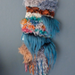 a woven wall hanging on a bamboo dowel rod with looped yarn in various greys, blues, greens, oranges, red and neutral browns too! gorgeous dark teal fiber inspired by waterfalls spill over the looped sections. 